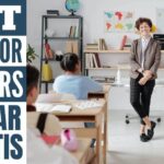 A female teacher standing in front of students in a class wearing glasses, a brown blazer, light blue shirt, dark grey & black checked pants with black shoes smiling